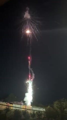 A nighttime video shows fireworks exploding in the sky. The fireworks are white and gold, and they create a burst of light and smoke. The video is taken from a low angle, and the fireworks are in the center of the frame. The sky is dark, and there are no other lights visible. The fireworks are a display of light and color against the dark sky.