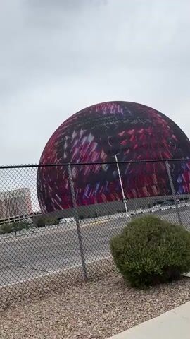 A video captures a large, spherical building covered in a digital display. The display shows a pattern of pink, purple, and red lights that appear to be moving. The building is partially obscured by a chain-link fence in the foreground. Behind the building, there are other structures and some vehicles on a road. The sky is overcast. Two small bushes are visible in front of the fence, and a trash receptacle is also present. A few bare tree branches are in the upper right corner of the frame.