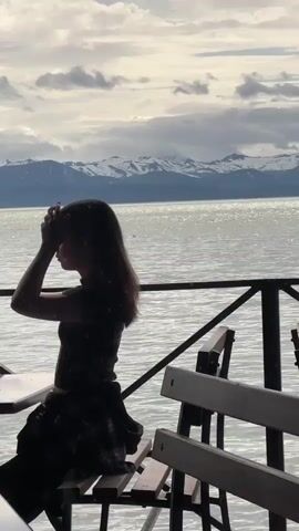 A young girl sits at a table by a railing, facing away from the camera, with her hands touching her head. Beyond the railing, a body of water reflects the sky and snow-capped mountains in the distance. The sky is cloudy, and the overall lighting suggests it is daytime. Wooden benches are visible in the foreground.