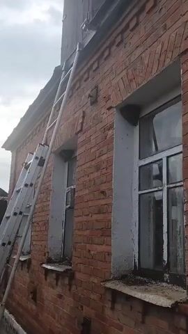 A silver ladder leans against the brick wall of a building, near two windows. The ladder extends to the top of the frame. The building has a wooden structure visible above the brickwork. The sky is overcast.