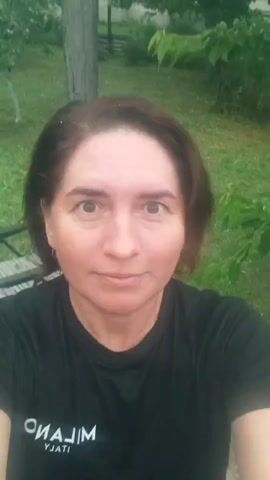 A medium shot video captures a woman with shoulder-length, reddish-brown hair wearing a black t-shirt. She is outdoors, with a blurred background of green foliage and trees, suggesting a natural setting. The lighting is somewhat diffuse, possibly indicating an overcast day or shade. The woman's face is the primary focus, and she appears to be looking towards the camera with a slight smile. Her head is tilted slightly to her right. The video is shaky, indicating it was likely filmed handheld. The woman's t-shirt has white lettering on it, with the word "MILANO" visible.