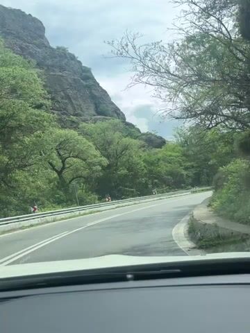 A car drives along a winding asphalt road. On either side of the road are lush green trees, vegetation, and rocky hills. A gray guardrail runs along one side of the road, and a low stone wall is on the other. The sky is partly cloudy with a mix of gray and white clouds. The view is from inside the car, looking through the windshield. Music is playing.
