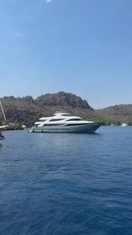 A large white yacht is anchored in the blue ocean, with green hills and a clear sky in the background. The yacht is the central focus of the video.