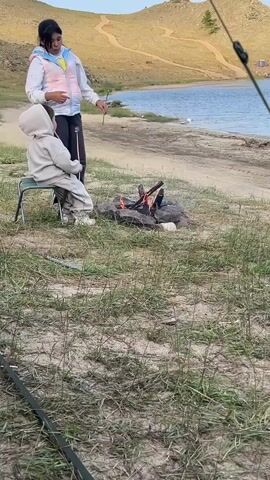 Two young females are gathered around a campfire on a sandy shore next to a body of water. The female on the left is seated on a metal folding chair, wearing a light gray hooded tracksuit. She holds a stick with a marshmallow over the fire. The female on the right is standing, wearing a puffy vest with multi-colored panels over a white top and dark pants. She holds a stick with a marshmallow, appearing to guide the other marshmallow over the flames. The campfire is built with rocks and burning logs. In the background, a body of water with calm waves is visible, and rolling, dry hills with winding paths rise behind the water. The sky is a pale blue, suggesting either early morning or late afternoon. The ground is covered in sparse green grass and sand.