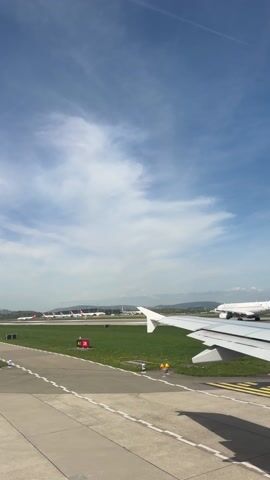 The video shows an airplane's perspective while taxiing on a runway. The sky is blue with scattered clouds. In the distance, several planes are parked, and hills can be seen. The plane's wing is visible in the foreground. Runway markings and signs are visible on the ground.