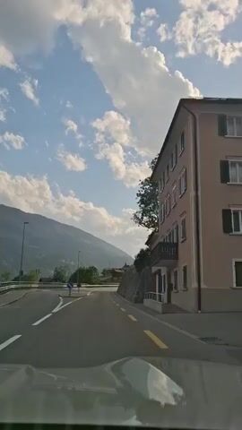 A vehicle is driving on a paved road. The road curves to the right and is lined with a stone wall on the right and a guardrail on the left. In the distance, there are mountains under a cloudy sky. A building with multiple windows and a balcony is visible on the right side of the road.