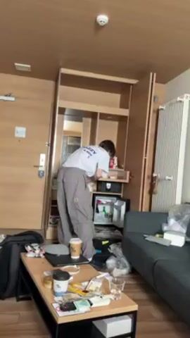 A person in a white t-shirt and gray pants is bending over a mini-fridge in a hotel room. The mini-fridge is built into a wooden cabinet. A coffee table in the foreground is cluttered with coffee cups, electronics, and other items. A gray sofa is visible to the right of the cabinet, and a suitcase is to the left. The room appears to be somewhat messy. The lighting is bright and even. The person is rummaging through the mini-fridge.
