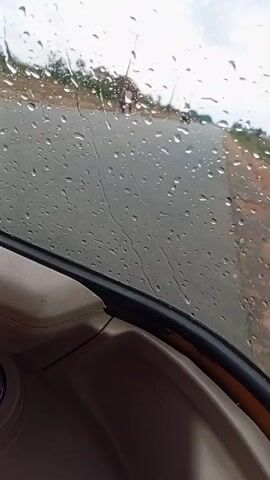 The view from inside a vehicle driving on a wet road shows raindrops obscuring the windshield. Other vehicles are visible on the road, and utility poles line the roadside. The interior of the vehicle has a light brown dashboard.  A QR code sticker is adhered to the lower corner of the windshield. The windshield wipers are in operation.
