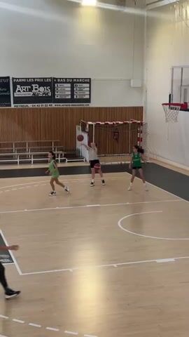 In an indoor basketball court in Rousset, France, several young females are engaged in a basketball activity. The court features a light wooden floor with white markings and a single basketball hoop. White walls with brown slatted panels and benches line the perimeter, along with a handball goal. Overhead, bright artificial lights illuminate the space.

The video captures a moment during practice or a casual game. One player, wearing a black top and shorts with pink shoes, shoots a basketball towards the hoop from near the free-throw line, successfully making the shot. Another player in a white t-shirt stands near the shooter, holding a ball. A player in a green jersey dribbles a basketball on the right side of the court, while another in a red t-shirt dribbles near the baseline. A fifth player, wearing a red vest over a dark long-sleeved top, walks slowly in the foreground.

Prominently displayed on the wall are several French advertisements. These include "LA CÔTE DE BOEUF" (The beef rib), "Boucherie Traditionnelle" (Traditional butcher shop), "PARMI LES PRIX LES PLUS BAS DU MARCHÉ" (Among the lowest prices on the market), "ART BOIS MENUISERIES ALU-PVC" (ART BOIS Aluminum-PVC joinery) with a phone number (04 42 51 27 48) and website (WWW.ARTBOIS-MENUISERIES.COM), and "GARAGE DE LA ROQUE". A scoreboard is also visible above the basketball hoop.