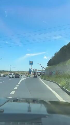 The video shows a driver's perspective of approaching a toll plaza on a highway. The sky is blue with a few wispy clouds. Power lines run overhead. The road is asphalt with white lane markings.  Several vehicles are visible in the distance. A large "AYGAZ" sign is visible on the left.  Turkish flags and a yellow and blue flag are displayed along the right side of the road. A concrete barrier runs along the right side of the road, covered with some graffiti. The hood of the car taking the video is visible at the bottom of the frame.