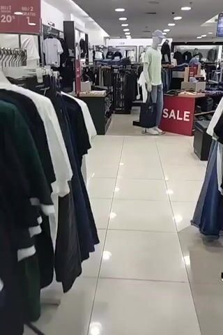 The video shows the interior of a clothing store. The camera pans across rows of clothing racks displaying various garments, including t-shirts, jeans, and jackets. Mannequins are visible, dressed in casual attire. Signs indicating sales and discounts are posted throughout the store. The lighting is bright, and the floor is tiled. A few customers can be seen browsing the merchandise in the background. The overall atmosphere appears to be that of a typical retail environment.