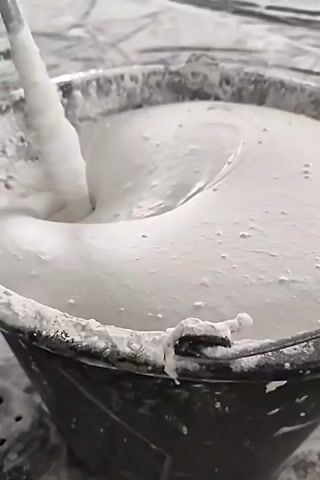 A close-up, overhead shot shows a grey mixing paddle stirring a thick, white, plaster-like substance in a black bucket. The plaster is being vigorously mixed, creating swirls and waves. The bucket is dirty with dried plaster around the rim. The background is out of focus and appears to be a grey, textured surface.