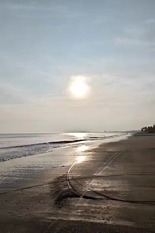 The scene is a beach on a sunny day. The sun is shining brightly, reflecting off the ocean and wet sand. The waves are small and gentle, lapping at the shore. The beach is wide and sandy, with some dark patches of wet sand. There are a few dark lines across the sand, possibly from equipment or vehicles. In the distance, there are some trees and buildings. There appears to be a person walking in the distance. The sky is a clear light blue with some scattered thin clouds.