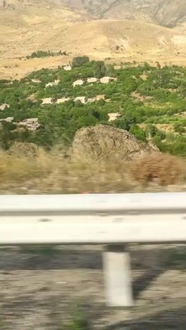 A blurry, horizontal video shows a landscape of dry, rocky hills and mountains. In the middle ground, there is a green patch of trees with several small buildings nestled within it. In the foreground, a white guardrail with metal posts is visible, indicating that the footage was likely filmed from a moving vehicle on a road. The sky is a pale blue with some light clouds. The overall impression is of a rural, arid environment.