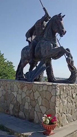 A bronze statue of Saint George on horseback is depicted, with the saint piercing a dragon with his spear. The statue is mounted on a stone pedestal. In the background, a landscape unfolds with mountains, a town, and green fields under a clear blue sky. The scene appears to be outdoors in daylight.