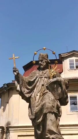 The video shows a statue of a man holding a cross and a palm branch, with a halo of stars around his head. The statue is made of stone and is located in front of a building with a red roof and white windows. The sky is blue and clear.