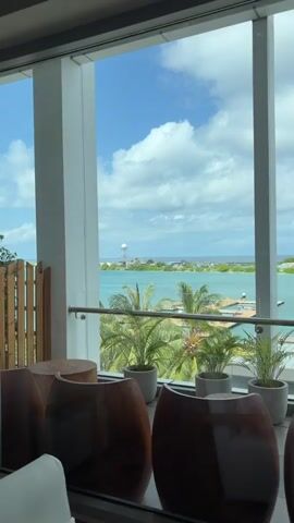 A video pans across a room with large windows overlooking a tropical ocean view. Inside, the room is furnished with modern, minimalist furniture, including white upholstered chairs, wooden coffee tables with natural wood tops, and a tall, woven floor lamp. Tropical plants are visible on the balcony outside. A framed picture of a sunset with curtains hangs on the wall. A white sheer curtain hangs from the ceiling to the floor on the right side of the frame. The lighting is soft, with the floor lamp casting a warm glow. The overall atmosphere is serene and luxurious.