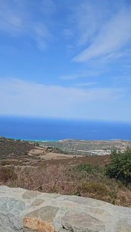 The video shows a panoramic view of a coastal landscape from a slightly elevated position. The foreground features a rough stone wall, partially overgrown with dry grasses and a few green plants. Beyond the wall, the terrain slopes down, revealing dry, scrubby vegetation and patches of cultivated land, possibly agricultural fields. Further in the distance, the vibrant blue sea stretches out to the horizon, meeting a bright blue sky dotted with wispy white clouds. The water near the coast appears a lighter turquoise, suggesting shallow areas or sandy seabeds. Scattered on the sea are a few small white specks that could be boats. A town or settlement with scattered buildings is visible along the coastline. The overall impression is of a sunny, dry, and scenic coastal environment.