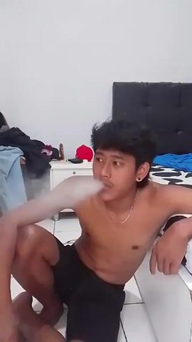 A shirtless young man is sitting on the floor in a bedroom. He is wearing black shorts and a gold chain. He has dark hair with a short mullet and a small earring in one ear. He is smoking a cigarette and exhaling smoke.  There is a bed behind him with a black headboard and a blue and white patterned blanket. To his left is a white nightstand with various items on it, including clothes, a red baseball cap, and a small object.  The walls are white, and the floor is tiled.