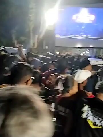 This is a crowd of people gathered at night watching a large outdoor screen. The screen displays a concert or performance, with bright lights and a stage visible. The people in the foreground are mostly facing the screen, and some are holding up phones, likely to record. The environment appears to be outdoors, possibly a park or open field, with trees visible in the background. The lighting is dim, with the screen being the main source of illumination. The crowd is dense, indicating a popular event.