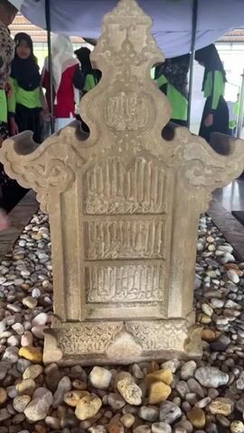 The video shows a tombstone with Arabic inscriptions, surrounded by small rocks. Several people are visible in the background, some wearing headscarves and traditional clothing, suggesting a religious or historical site. The tombstone is made of light-colored stone and has ornate carvings at the top and base. The overall scene appears to be outdoors, possibly under a tent or canopy, indicating an event or a tourist visit. The lighting is natural, suggesting daytime.
