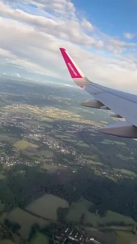 The video captures a view from an airplane window, showcasing the landscape below during flight. The prominent subject is the wing of an aircraft, with the Wizz Air logo and "wizzair.com" clearly visible on its pink wingtip. The scene depicts a sprawling, verdant landscape consisting of a patchwork of green fields, dense forests, and scattered residential areas or towns. Several buildings with white roofs are visible among the trees and fields, suggesting human habitation. In the distance, wind turbines can be faintly discerned against the horizon. The sky above is partially cloudy, featuring white cumulus clouds against a backdrop of clear blue. The lighting suggests it is daytime, possibly late afternoon or early evening. The aircraft appears to be at a relatively low altitude, allowing for detailed observation of the ground. Given the provided context, the flight is likely taking place over Holzwickede, Germany. The video is static, focusing on the scenic aerial view, and includes the ambient sound of the airplane.