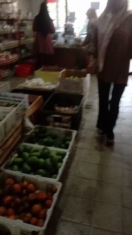 The video shows an indoor scene with a person walking through what appears to be a market or store. The person is wearing dark pants and a patterned top. There are shelves with items on them to the left, and produce displayed in baskets or crates to the right. The lighting is somewhat dim. The camera is moving and the footage is shaky.