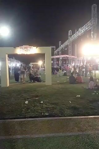 The video begins with a view of an outdoor event at night. In the foreground, there is a grassy area with some litter. In the mid-ground, there is a large, archway structure with the words "MEMETRI 2023" visible on it. Several people are standing near and within the archway, some of them appearing to be interacting. To the left of the archway, there are white tents. In the background, there is a stage or lighting rig with bright lights and other structures associated with an event. There are more people scattered throughout the background, some seated and others standing. The lighting is artificial, with bright spotlights and ambient illumination from the tents and stage area. The video then pans to the right, showing a glimpse of what appears to be a track with colored lanes. The overall impression is of a lively, open-air event taking place at night.