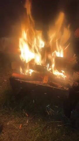 This is a close-up video of a campfire at night. The flames are bright orange and yellow, and they flicker and dance in the darkness. The fire is contained within a metal grill, and there are logs burning inside. The ground around the fire is covered in grass, and there are several cigarette butts scattered on the ground. In the background, there are faint outlines of people, suggesting a gathering around the fire. The overall atmosphere is cozy and warm, despite the darkness.