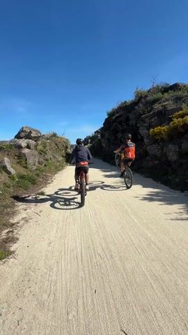Two people are riding bicycles on a dirt path. The person in front is doing a wheelie. Both people are wearing helmets and athletic clothing. The path is bordered by rocks and foliage on either side. The sky is clear and blue.