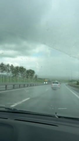 The video shows a view from inside a car driving on a highway. The windshield is dirty and wet, suggesting rain. The road is wet and appears to have standing water in some areas. Several cars and a truck are visible on the road ahead, as well as to the side. The sky is overcast with dark clouds. Trees and greenery are visible along the side of the highway. The video is shaky, indicating movement.
