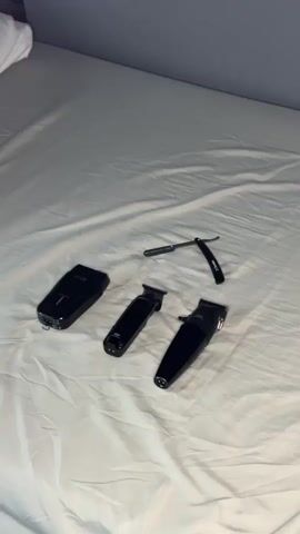 A white comforter covers a bed in a room with a gray wall. Four grooming objects are on the comforter. On the left is an electric razor. To the right of the razor are two electric trimmers. All three of the electrical items are black. To the right of the trimmers is a straight razor. Someone sprays the items with a clear substance from an unknown source.