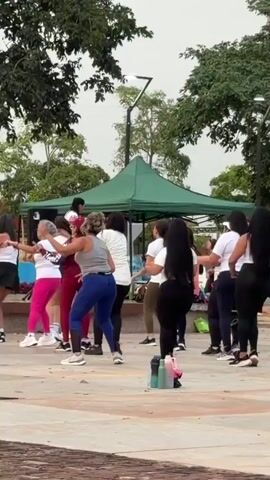A group of women are engaged in a dance fitness class outdoors. They are positioned in a line facing to the right of the frame, with several individuals also visible behind them. The women are dressed in athletic wear, primarily white tops and dark-colored leggings or pants, with one woman in red leggings and another in blue leggings. They are all performing synchronized dance moves, with arms extended and bodies in motion. A green tent is situated in the background, and trees and streetlights are also visible, suggesting an urban park setting. Several water bottles are placed on the ground in the foreground. The overall atmosphere appears energetic and recreational.
