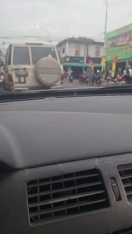 The video, taken from inside a car, shows a street scene on a cloudy day. In front of the car is a light-colored SUV with a spare tire mounted on its back. Several motorcycles are parked or moving along the street. Buildings with shops and restaurants line the street, with people visible near the entrances. A flag is visible in the background. The car's dashboard is visible in the foreground, including air vents and a slightly cracked surface.