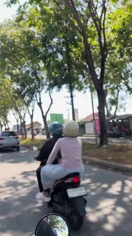 The video shows a rear view of a motorcycle with two people riding on it. The driver is wearing a dark jacket and a blue helmet. The passenger is wearing a light pink shirt and a light-colored helmet, and they are sitting behind the driver, facing forward. The motorcycle is moving along a paved road, with trees lining the side. There are other vehicles and buildings visible in the background. The overall scene appears to be a sunny day.