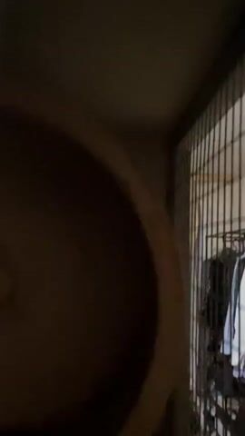 This is a video clip showing a chinchilla inside a multi-level cage. The chinchilla is dark gray with lighter markings and has large ears. It is perched on a shelf, looking down at the camera. The cage appears to be made of metal bars and has wooden platforms. The lighting is dim, suggesting it may be nighttime or indoors with low light. The background is mostly out of focus, but hints of the surrounding room can be seen, including what looks like a wooden structure and more cage components. There is no visible text in the clip. The activity is simply the chinchilla existing within its habitat, with a focus on its presence and appearance. The setting is indoors, likely a home environment, given the domestic nature of the animal and its enclosure. The chinchilla's posture and gaze suggest curiosity or perhaps a slight wariness of the camera.