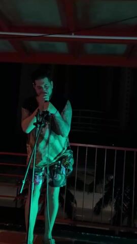 Here is a description of the video:
A man is singing into a microphone on an outdoor stage at night. He has tattoos on his arms and is wearing shorts and a sleeveless shirt. The lighting changes from green to red. The stage has a railing and a partial roof overhead. In the background, there are palm trees, lights from buildings and streetlights.