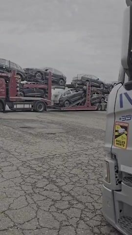 The video shows a car carrier truck loaded with several cars. The truck is red and appears to be a Scania model, with "SCANIA" visible on the front grille. The trailer is a multi-level rack designed to transport multiple vehicles. The cars on the truck are dark colored, likely black or dark grey, and appear to be sedans or similar passenger vehicles. Some are on the top level, and others are on the lower levels of the trailer.

The truck is parked on a paved area with visible cracks in the surface, suggesting it might be a lot or a storage yard. In the background, there is a green corrugated metal fence and a concrete wall. A street light or lamp post is also visible in the distance. The sky is overcast, indicating a cloudy day. The overall scene appears to be an outdoor industrial or logistics area. There is no visible human activity.

The text "AUTO" is visible on the side of the truck's cab. The timestamp at the beginning of the video shows "00:00", followed by subsequent timestamps indicating the duration of the clip. The location context provided is Épila, Spain.