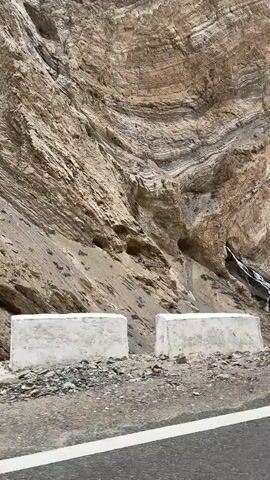The video shows a mountainous landscape with a large rock face. The rock face is layered and appears to be made of various shades of brown and gray rock. There is a small waterfall cascading down a section of the rock face. In the foreground, there is a paved road with white markings. There are also some concrete barriers on the side of the road. A small brown animal, possibly a goat or a sheep, is standing near the road. The overall environment appears to be arid and mountainous.