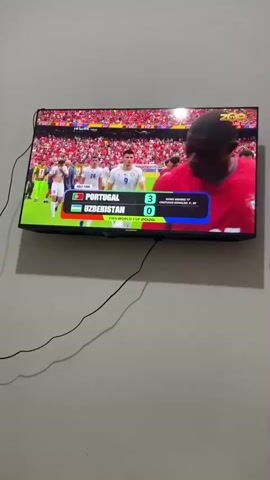 The video displays a television screen showing a soccer match between Portugal and Uzbekistan. The score is 3-0 in favor of Portugal. The match appears to be part of the FIFA World Cup 2026. The scene is bright, suggesting daylight, and the setting is indoors, likely a living room. The atmosphere seems to be one of watching a sporting event, with the focus on the game's progress. The screen displays player numbers and flags of the respective countries, indicating the teams involved. Following the soccer match, the screen transitions to a colorful animated scene featuring the Statue of Liberty and confetti, followed by the FIFA World Cup 2026 logo against a backdrop of a blue sky with clouds.