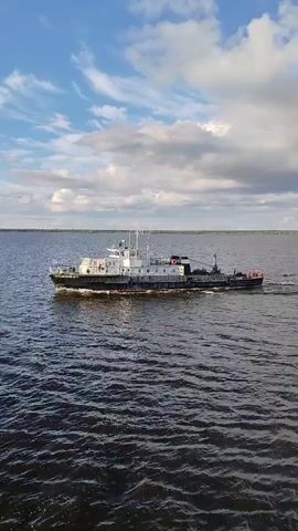 An aerial drone shot shows a white and gray ship moving across dark blue water under a partly cloudy sky. The ship appears to be traveling from left to right in the frame, leaving a wake behind it. The sky is a bright blue with scattered white clouds. The horizon line is visible in the distance, with land or a distant shoreline appearing as a faint, darker line. The overall impression is a serene scene of a vessel on a large body of water.