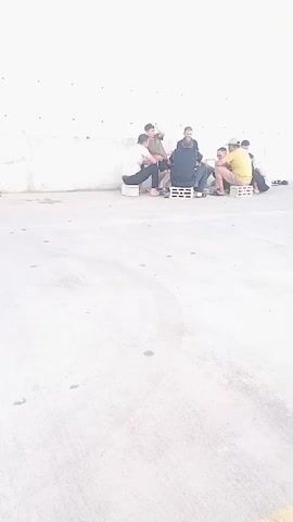 A group of eight individuals are gathered in a semi-circle against a plain white wall. The ground is concrete. The individuals are seated on a combination of wooden pallets and what appear to be small plastic crates. Most of the individuals are men, dressed in casual clothing. Some are interacting with each other, while others are looking down or away. The lighting suggests it is daytime.