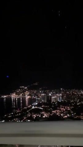 The video is a nighttime aerial view of a coastal city illuminated by numerous lights. The camera appears to be moving, possibly from a vehicle or drone, as the perspective shifts slightly. The city lights reflect on the dark water of a bay or ocean. In the foreground, a blurred railing or edge of a structure is visible. The sky is dark, with a few faint streaks of light, possibly distant lights or a lens flare.