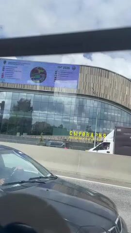 The video, shot from a moving vehicle, depicts the exterior of a building in Moscow, Russia, identified as "Syrovarnya" (Cheese Factory). The building has a modern, curved design with a glass facade and a textured wooden-like exterior. A large ISF 2026 banner is visible on the building's roof. Traffic flows on the road in front of the building. The sky is partly cloudy.