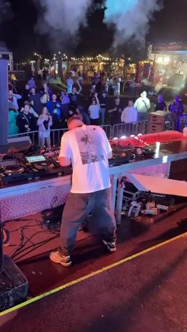 The video shows a DJ performing at an outdoor event at night. The DJ, a man with short blond hair wearing a white t-shirt and grey pants, is standing behind a DJ booth with turntables and other equipment. He is looking down at the equipment and appears to be mixing music. A crowd of people is gathered in front of the DJ booth, some of whom are dancing and raising their hands. There are lights and smoke effects in the background, suggesting a lively atmosphere. The scene is lit by stage lighting and string lights in the background trees. There is a barrier in front of the DJ booth separating the DJ from the crowd. The event appears to be taking place in a public space, possibly a park or a street, given the presence of trees and buildings in the background.