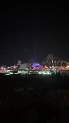 The video shows a nighttime scene with a large cable-stayed bridge illuminated in the background. To the left of the bridge, there is a Ferris wheel and what appears to be a large building complex. A portion of the building complex is lit with purple lights, and there are beams of light projecting upwards from it. In front of the buildings, there are various lights from what seem to be vehicles and smaller structures, suggesting an urban or industrial area. The foreground shows the dark silhouettes of trees and foliage. The sky is dark, indicating it is nighttime, with a few faint specks that could be distant lights or atmospheric phenomena. The overall impression is a vibrant, illuminated cityscape or event taking place at night, with the bridge serving as a prominent landmark.