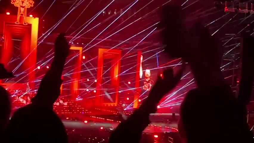 The video shows a concert or live performance. The stage is illuminated with bright red lights and beams. The audience is visible in the foreground, with their silhouettes raised in the air, some with hands in the air, indicating engagement and excitement. The stage has large, rectangular, illuminated structures that appear to be part of the set design. A figure, likely the performer, is visible on the stage in the distance, also illuminated by the stage lights. The overall atmosphere is energetic and festive.
