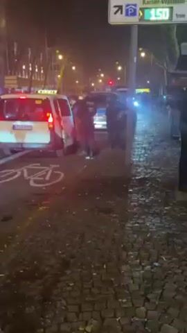 The video shows a street scene at night, likely in a European city. Several police officers are standing near police vehicles, including a van with the word "POLIZEI" and "NRW" (likely referring to Nordrhein-Westfalen, a German state) on the back. The ground is littered with debris. Other people are walking around and standing on the sidewalk near a building identified as "Inception Hotel." Bicycles are parked nearby. The scene suggests some sort of public disturbance or police presence following an event.