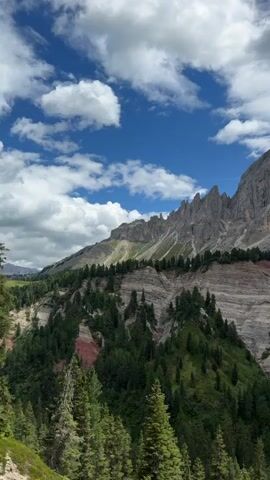 This video shows a scenic view of a mountain range with a forest in the foreground. The mountains are steep and rocky, with some snow visible on the peaks. The forest is dense with evergreen trees. The sky is blue with some white clouds. The video appears to be shot from a high vantage point, possibly from a hiking trail or a helicopter. There are no people or animals visible in the video. The environment is natural and undeveloped.