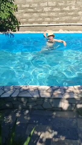 A person wearing a straw hat is swimming in a pool. They are in the center of the pool and appear to be moving their arms. The water is blue and sunny, with ripples on the surface. The pool is surrounded by a tiled border and a brick wall. In the foreground, there are some plants. Another person, a child, is partially visible in the pool on the right side of the frame, near the edge.
